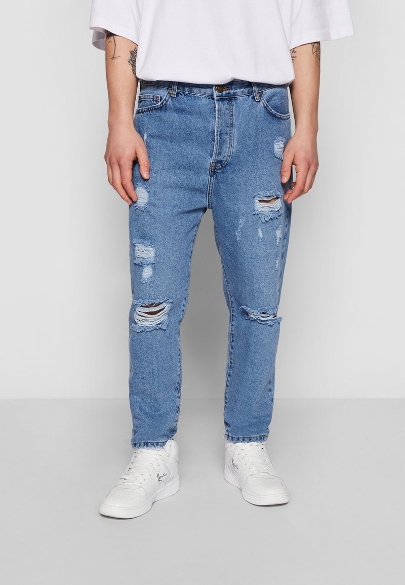Personne portant un jean bleu clair déchiré, des baskets blanches et une chemise blanche à manches courtes, debout devant un fond uni.