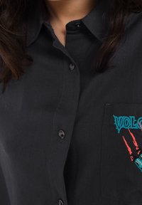 Chemise noire à boutons avec une poche, ornée de motifs de griffes brodés en bleu et rouge ainsi que du mot "VOLCANO". Tissu en coton avec une texture lisse.