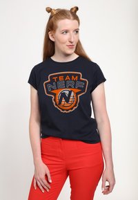 Námornická tričko s grafikou "TEAM NERF" v oranžové a bílé. Krátké rukávy, kulatý výstřih, uvolněný střih. Viditelné červené kalhoty.