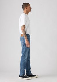 Hvit bomull T-skjorte, blå denim jeans, svarte joggesko med hvite såler. Personen står i profil mot en ensfarget bakgrunn.