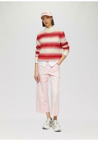 Strickpullover mit roten und cremefarbenen Streifen, kombiniert mit hellrosa weit geschnittenen Hosen. Das Modell trägt eine rosa Kappe und graue Sneakers. Strukturierte Ausführung.