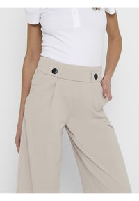 Frau trägt beigen hoch-taillierten, weit geschnittenen Hosen mit schwarzen Knöpfen und einem weißen, gerippten Kurzarmoberteil, mit der Hand in der Tasche, vor einem einfarbigen Hintergrund.