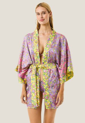 Donna che indossa un kimono floreale colorato con bordo giallo legato in vita sopra un bikini abbinato su sfondo semplice.