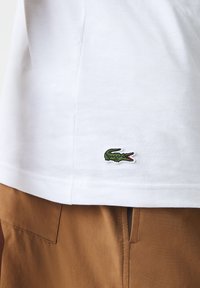 Bílý bavlněný tričko s vyšitým zeleným logem krokodýla na lemu. Jednoduchý design s klasickým kulatým výstřihem.