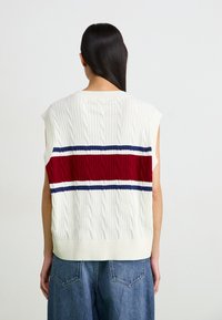 Brezrokav pleteni pulover v slonokoščeni barvi z mrežastim vzorcem, ki ima na hrbtu širok rdeč trak, obdan z mornarsko modro in belo.
