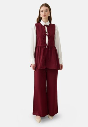 Donna che indossa pantaloni bordeaux a gamba larga e tunica senza maniche abbinata sopra una camicia bianca a maniche lunghe con tacchi beige.