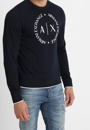 Mężczyzna noszący czarną bluzę Armani Exchange z długim rękawem i białym logo, w połączeniu z postrzępionymi niebieskimi dżinsami.