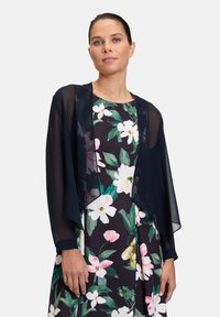 Schwarzer, transparenter Umhang über einem floral gemusterten Kleid mit dunklem Hintergrund. Mit weiten Ärmeln, subtiler Schattierung und schichtigem, fließendem Design.