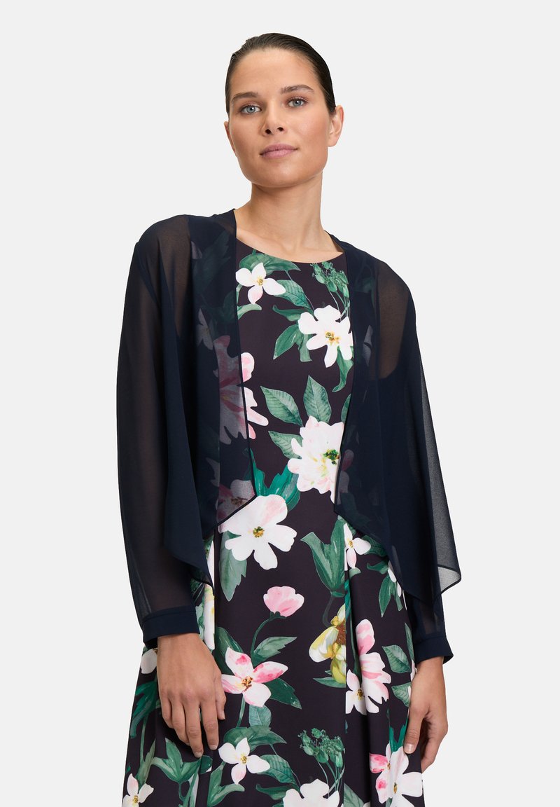 Schwarzer, transparenter Umhang über einem floral gemusterten Kleid mit dunklem Hintergrund. Mit weiten Ärmeln, subtiler Schattierung und schichtigem, fließendem Design.