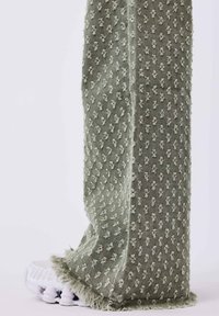 Pantalón de tejido verde con un patrón, superficie texturizada y dobladillo deshilachado, combinado con una zapatilla deportiva blanca.