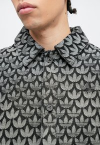 Artinys žmogaus, dėvinčio užsegamą pilkos ir juodos spalvų striukę su pasikartojančiu lapų raštu ir apykakle.