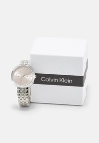 Sølv rustfritt stålklokke med en rund, lys rosa urskive og tre sub-dialer, ved siden av en hvit boks med en svart stropp med "Calvin Klein" tekst.