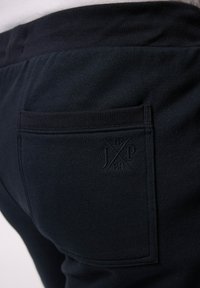 Mörknavy joggers med en ribbad midja och bakficka, texturerat tyg med en präglad logotyp som läser "JXP 18/80" på fickan.