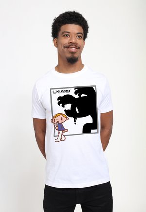 Un homme souriant porte un T-shirt blanc avec un dessin animé d'un enfant léchant une glace et une grande silhouette noire et ombragée de monstre derrière.