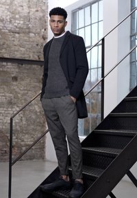 Abrigo negro sobre un suéter de canalé gris, combinado con pantalones grises con patrón y zapatillas negras. El modelo está de pie en una escalera de metal.