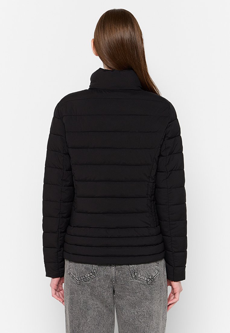 Giacca puffer nera con trapuntatura orizzontale, colletto alto e design aderente, abbinata a jeans grigi chiari strappati.