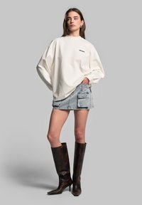 Witte oversized langekleurige top met lange mouwen, strakke denim mini-skirt met zakken, en hoge bruine leren puntige laarzen. Eenvoudige, casual stijl.