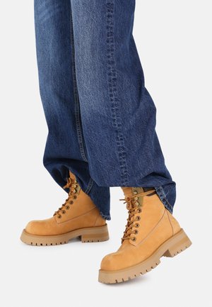 Botas de ante color tostado con un diseño robusto, atadas con cordones, que presentan una suela gruesa y texturizada y ojales de metal, combinadas con unos jeans de mezclilla azul sueltos.