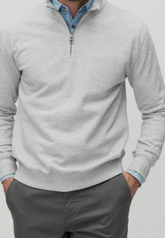 Homme portant un pull gris clair à fermeture éclair quart de zip sur une chemise bleue à boutons, les mains dans les poches d’un pantalon gris foncé, cadré au niveau du cou et des cuisses.