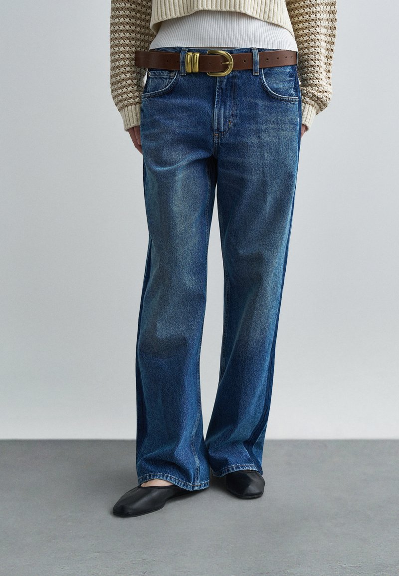 Blugi albastri cu talie largă, din denim, cu un aspect deschis la culoare, design cu cinci buzunare și curea maro din piele cu cataramă aurie. Pantofi negri comozi vizibili.