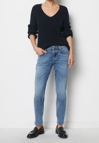 Person klädd i en mörk marinblå V-ringad tröja, ljusblå högmidjade jeans med dubbla knappar och svarta läder loafers, står mot en vit vägg.