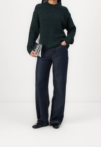 Dunkelgrüner, grob gestrickter Pullover mit geripptem Muster, kombiniert mit dunkelblauen Jeans mit weitem Bein. Silberne Clutch und schwarze Schuhe runden das Outfit ab.