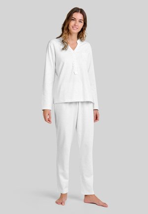 Mujer de pie descalza, vistiendo una blusa blanca de manga larga con botones y pantalones blancos a juego, frente a un fondo gris liso.