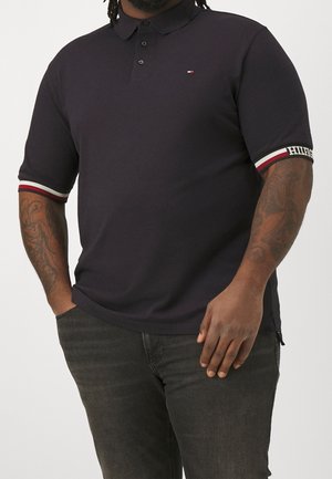 Homme portant un polo noir à manches courtes avec des poignets rayés rouge, blanc et noir et un jean foncé.