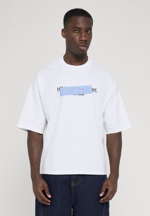 Ung mand iført en hvid oversized t-shirt med delvist tilsløret sort og blåt teksttryk, kombineret med mørkeblå jeans, mod en ensfarvet baggrund.