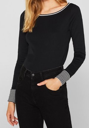 Kvinde iført en sort langærmet top med hvide og sorte stribede manchetter og halsudskæring, kombineret med sorte jeans med høj talje.