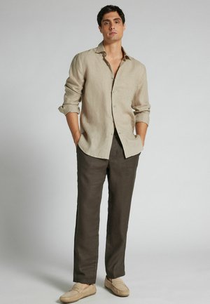 Młody mężczyzna ubrany w beżową lnianą koszulę z podwiniętymi rękawami, ciemnobrązowe spodnie i beżowe mokasyny, stojący z rękami w kieszeniach na jednolitym tle.