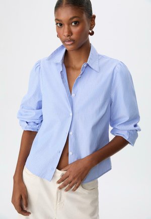 Donna che indossa una camicia leggera a righe blu chiaro con le maniche arrotolate e pantaloni color crema, davanti a uno sfondo bianco uniforme.