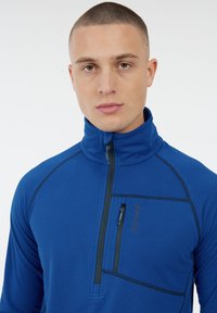 Elektrický modrý pulóver vyrobený z textúrovanej tkaniny. Má vysoký golier, polovičný zips a náprsné vrecko so zipsom. Čierne stehy ako akcenty.