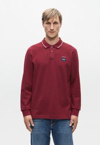 Bordeaux langemouwen polo met een klassieke kraag, witte bies en een klein blauw logo-opdruk op de borst. Gekocht met blauwe spijkerbroek.
