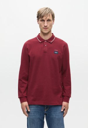 Burgundy long-sleeve polo shirt with a classic collar, white piping, and a small blue logo patch on the chest. Worn with blue jeans.