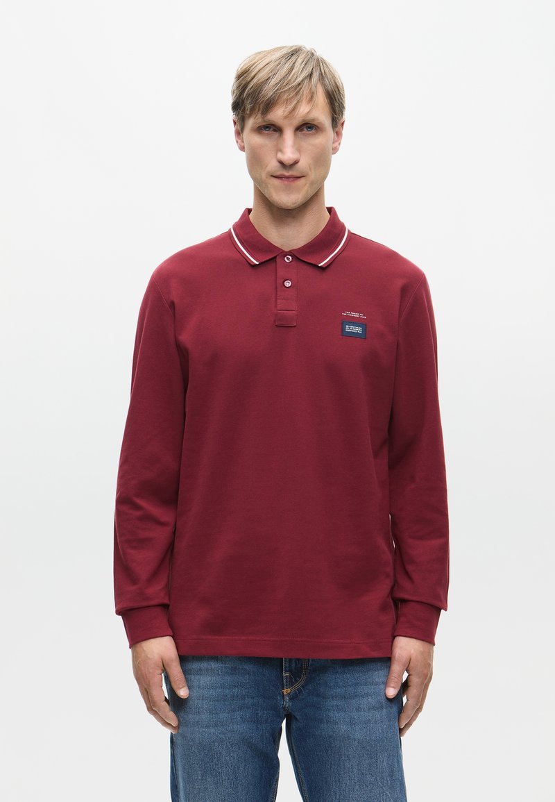Bordeaux langemouwen polo met een klassieke kraag, witte bies en een klein blauw logo-opdruk op de borst. Gekocht met blauwe spijkerbroek.