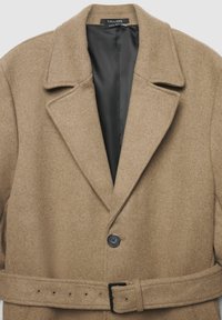 Cappotto in misto lana color marrone chiaro con colletto a risvolto, chiusura con un bottone singolo e una cintura abbinata in vita. Fodera nera visibile.