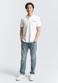 Weißes Poloshirt mit subtiler Textur, ausgestattet mit einem Druckdesign auf der linken Brust, kombiniert mit hellblauen Jeans und weißen Sneakern.