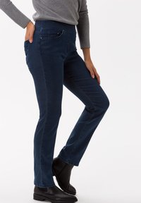 Pantaloni in denim blu scuro con vestibilità slim, caratterizzati da un ampio elastico in vita, tasche laterali e una texture liscia. Abbinati a stivaletti neri alla caviglia.