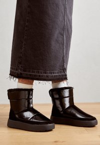 Svarta, glänsande, puffiga ankelstövlar med en mjuk pälsfoder och en tjock, texturerad sula; har två horisontella band över ovandelen.