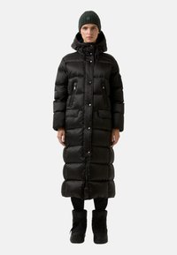 Schwarze Daunenjacke mit knielangem Design, Reißverschlusstaschen und Kapuze mit Kordelzug. Kombiniert mit schwarzen Stiefeln und einer grünen Strickmütze.