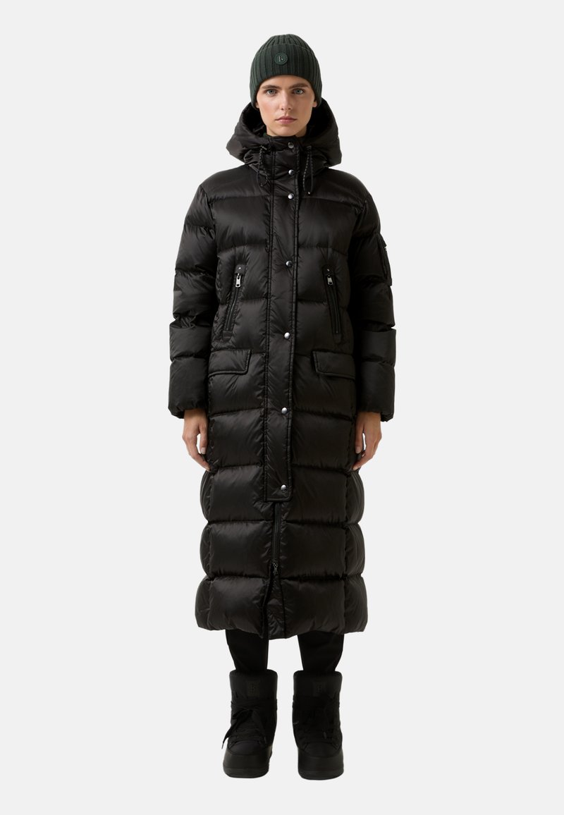Schwarze Daunenjacke mit knielangem Design, Reißverschlusstaschen und Kapuze mit Kordelzug. Kombiniert mit schwarzen Stiefeln und einer grünen Strickmütze.
