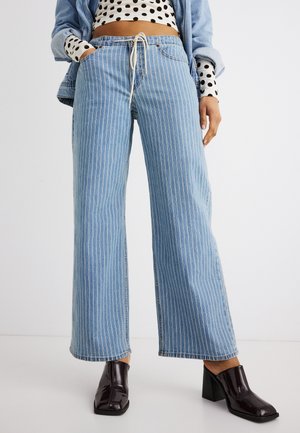 Frau trägt hochtaillierte blaue Denim-Weitbein-Hose mit weißen vertikalen Streifen, kombiniert mit schwarzen Loafers mit Absatz und einem gepunkteten Oberteil.