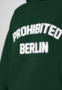 Sweat à capuche vert foncé avec un texte blanc en relief et en gras indiquant "PROHIBITED BERLIN" sur la poitrine et une poche avant.