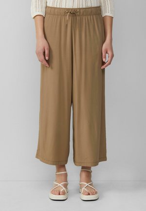 Pantalons larges marron avec une taille élastique et un cordon de serrage. Tissu doux, design sans embellissements, associés à des sandales blanches à brides.