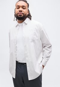 Άνδρας με γενειάδα και dreadlocks που φοράει λευκό πουκάμισο με ρίγες πάνω από ένα λευκό μπλουζάκι και σκούρο παντελόνι, στέκεται με το ένα χέρι στην τσέπη.