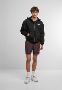 Felpa nera con zip, tasche frontali e dettagli del logo rosa, abbinata a pantaloni corti viola scuro e sneakers bianche, in piedi su uno sfondo grigio.
