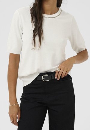 Kvinde iført en hvid kortærmet sweater med sort syning ved halsen, sorte bukser, et sort bælte og sølvringe på fingrene.