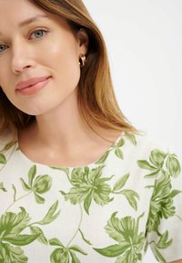 Lniany top z zielonym florystycznym wzorem, okrągłym dekoltem, krótkimi rękawami i gładką fakturą. Złote kolczyki w kształcie kół podkreślają całość.