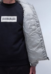 Vadderad grå jacka med glänsande yta, med dragkedja och elastiska ärmmuddar, bärs över en mörkblå sweatshirt med vit logotyp.